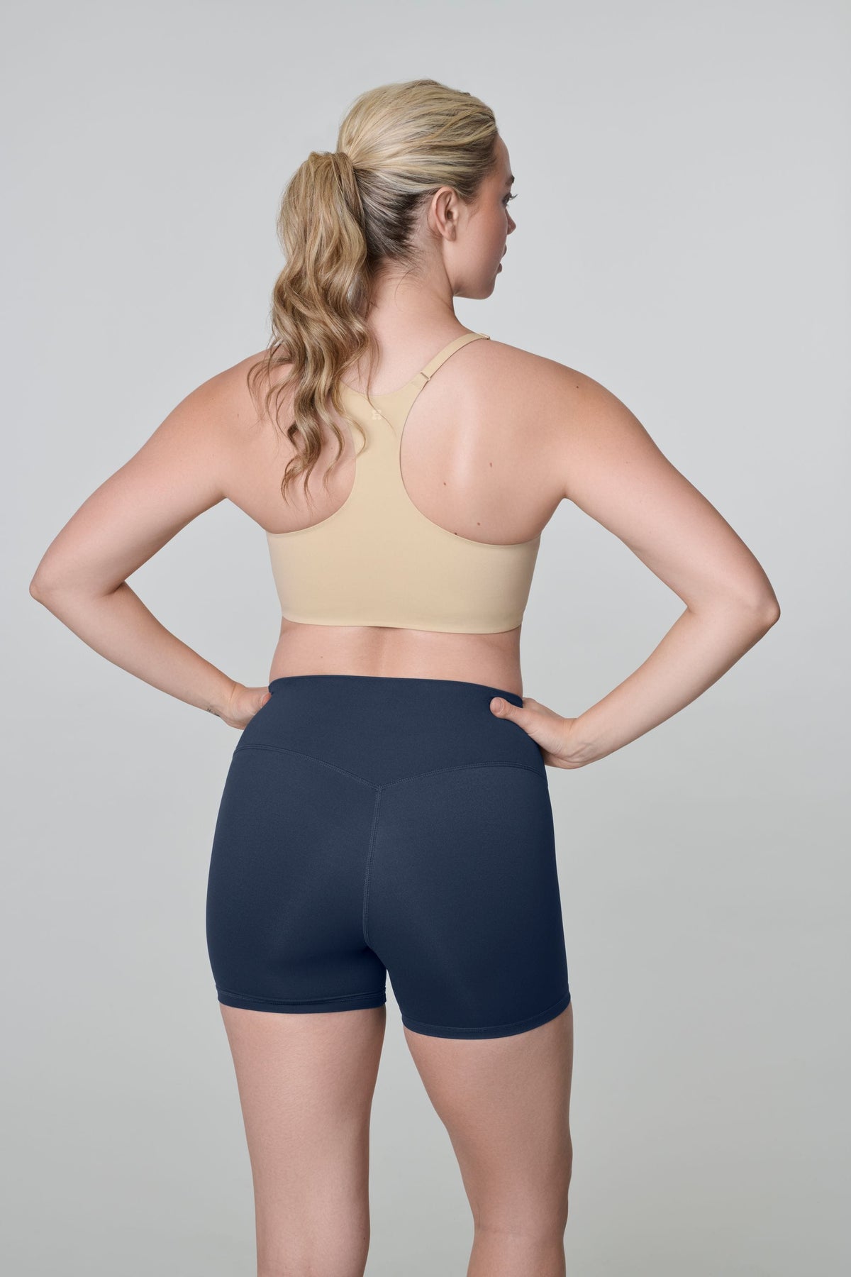 Bike Short in Navy, back view worn by a woman with a blonde ponytail and hands on her hips, with a nude racerback bra
